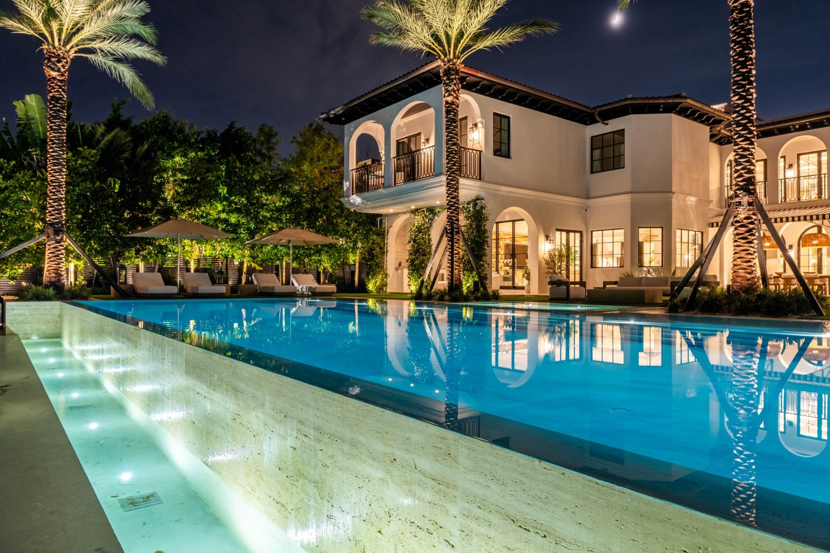 Long pool with travertine edge reflecting a Mediterranean-style home at night